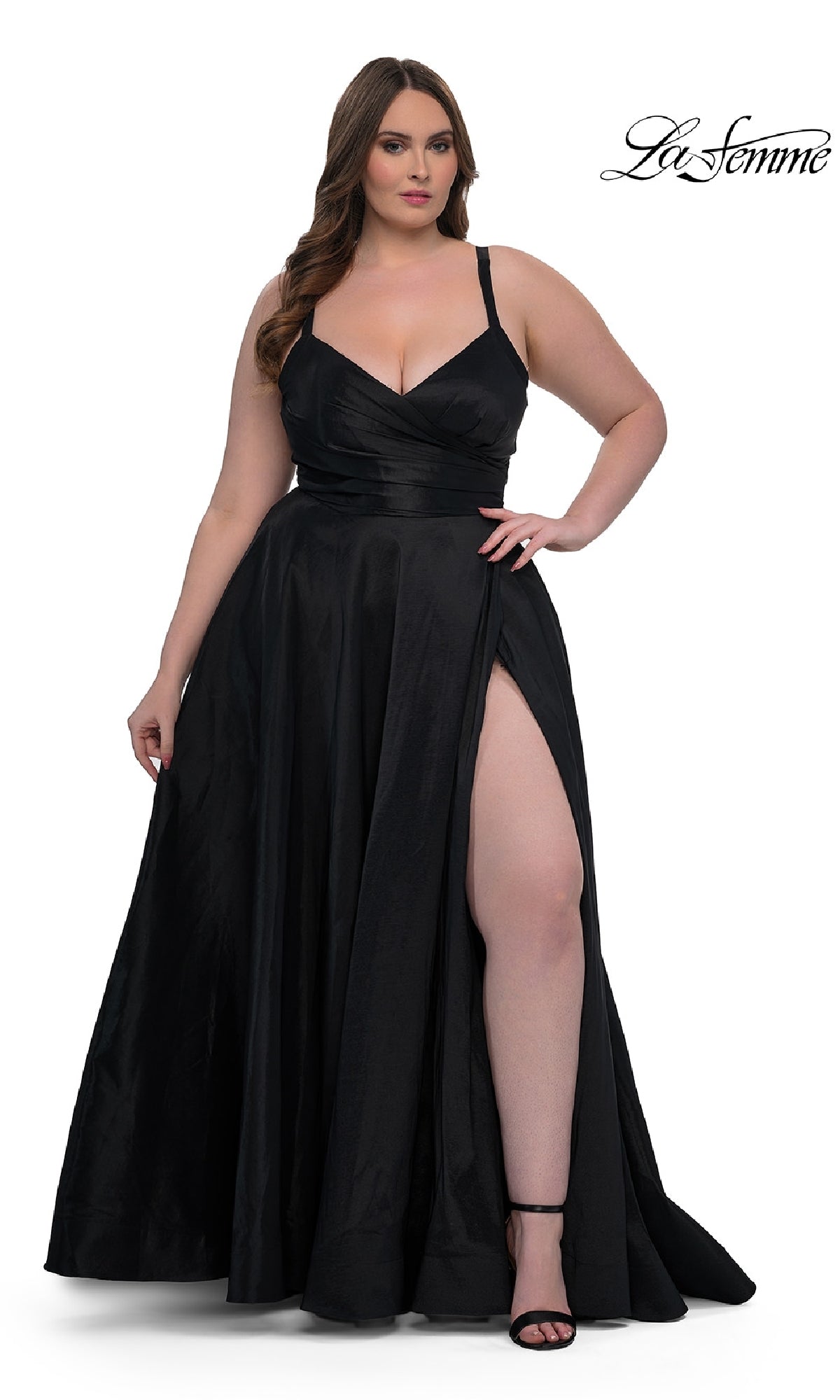 A woman stands against a white background in the La Femme 33075 long plus-size prom dress—black, sleeveless, with a faux-wrap v-neck and thigh-high slit—paired with black heels. The La Femme logo appears in the top right corner.